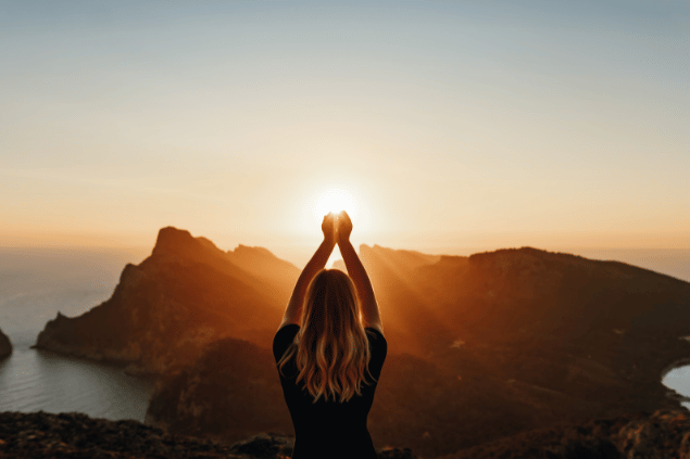 DianaHirsch / Getty Images Signature / Canva Uma mulher está com os braços levantados para o pôr do sol em uma praia.
