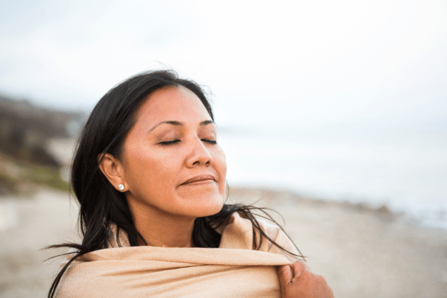 Uma mulher está em uma praia. Ela levanta a cabeça, fecha os olhos e sente a natureza.