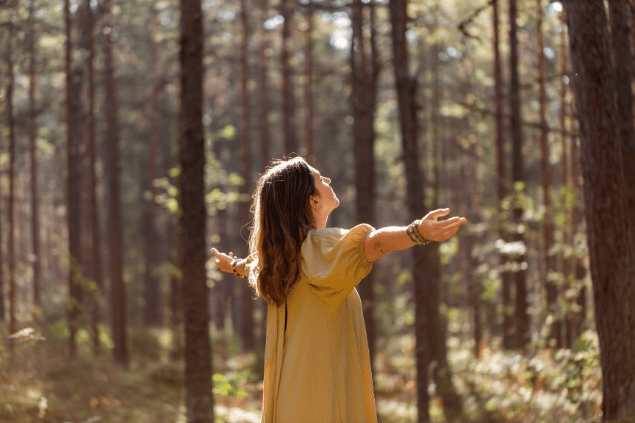 Uma mulher está com os braços abertos em uma floresta. Ela levanta a cabeça, fecha os olhos e sente os raios de sol na pele.
