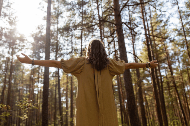Syda Productions / Canva Uma mulher está com os braços abertos em uma floresta. Ela levanta a cabeça e sente os raios de sol na pele.