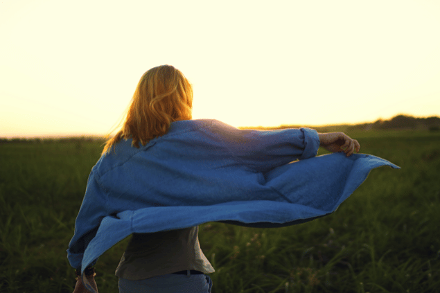 Fabriziogaleano10 / Pixabay / Canva Uma mulher está com os braços abertos em um ambiente de natureza. Ela segura sua camisa esvoaçante e sente os raios de sol na pele.