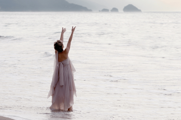 Barsik / Getty Images / Canva Uma mulher está de pé no raso do mar. Ela levanta a cabeça e os braços e sente a natureza, a espiritualidade.