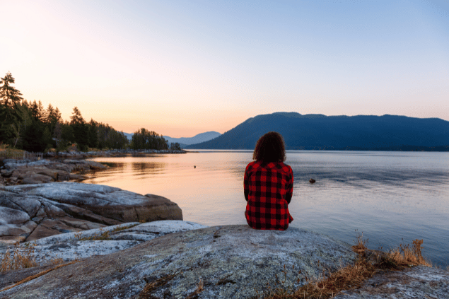 Edgar Bullon's Images / Canva Uma mulher está sentada na frente de um lago em uma floresta. Ela contempla e observa o pôr do sol.