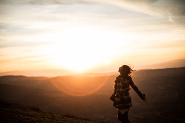 Miodrag ignjatovic / Getty Images Signature / Canva Uma mulher está com os braços abertos em um ambiente de natureza. Ela levanta a cabeça e sente os raios de sol na pele.