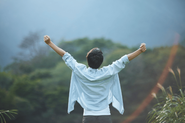 Aflo Images / アフロ(Aflo)/ Canva Um homem está com os braços levantados em um ambiente aberto. Ele levanta a cabeça e sente a natureza.
