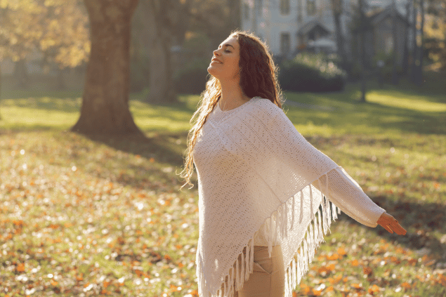 SrdjanPav / Getty Images Signature / Canva Uma mulher está com os braços abertos em um ambiente de natureza. Ela levanta a cabeça, fecha os olhos e sente os raios de sol na pele.