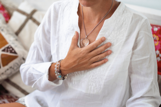 Microgen / Getty Images / Canva Uma mulher está com a mão no peito em sinal de paz, meditação. Ela usa roupa branca e um colar de cristal.