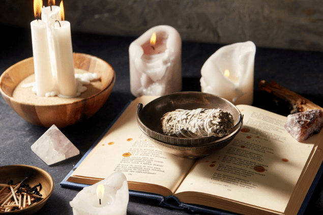 Syda Productions / Canva Na imagem, há alguns recipientes com sálvia branca, velas e fósforos em cima de uma mesa. Além disso, há cristais e um livro aberto.