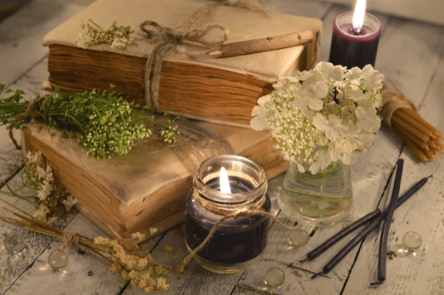 Na imagem, há alguns recipientes com flores e velas em cima de uma mesa. Há dois livros também.
