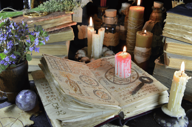 Vera Petruk / Canva Na imagem, há um livro aberto, velas acesas, livros fechados, um cristal e flores em cima de uma mesa.