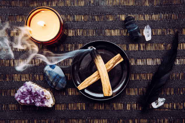 FotoHelin Images / Canva Na imagem, há um recipiente com palo santo em cima de uma mesa. Há uma vela, pedras e uma pena.
