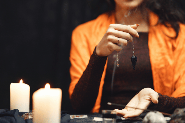 Gustavo Fring / Pexels / Canva Na imagem, as mãos de uma taróloga segurando um pingente com um cristal estão em destaque. Ao lado, há velas e cartas de Tarot.