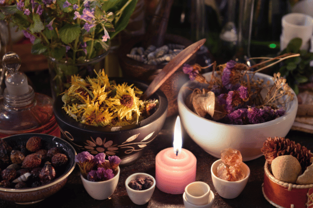 Na imagem, há alguns recipientes com flores e pedras em cima de uma mesa. E há uma  vela acesa.