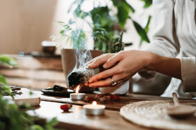 Nina_gili / Canva Na imagem, as mãos de uma mulher queimando uma sálvia branca estão em destaque. Há flores e velas ao lado.