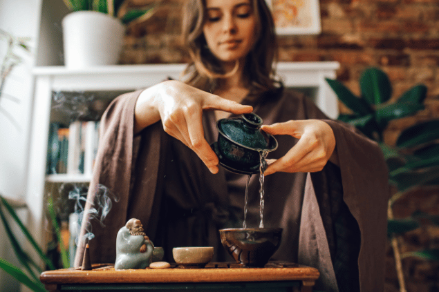 Roman Odintsov / Pexels / Canva Na imagem, as mãos de uma mulher enchendo um recipiente com água estão em destaque. Há um incenso, uma estátua e uma pedra em cima de uma mesa.