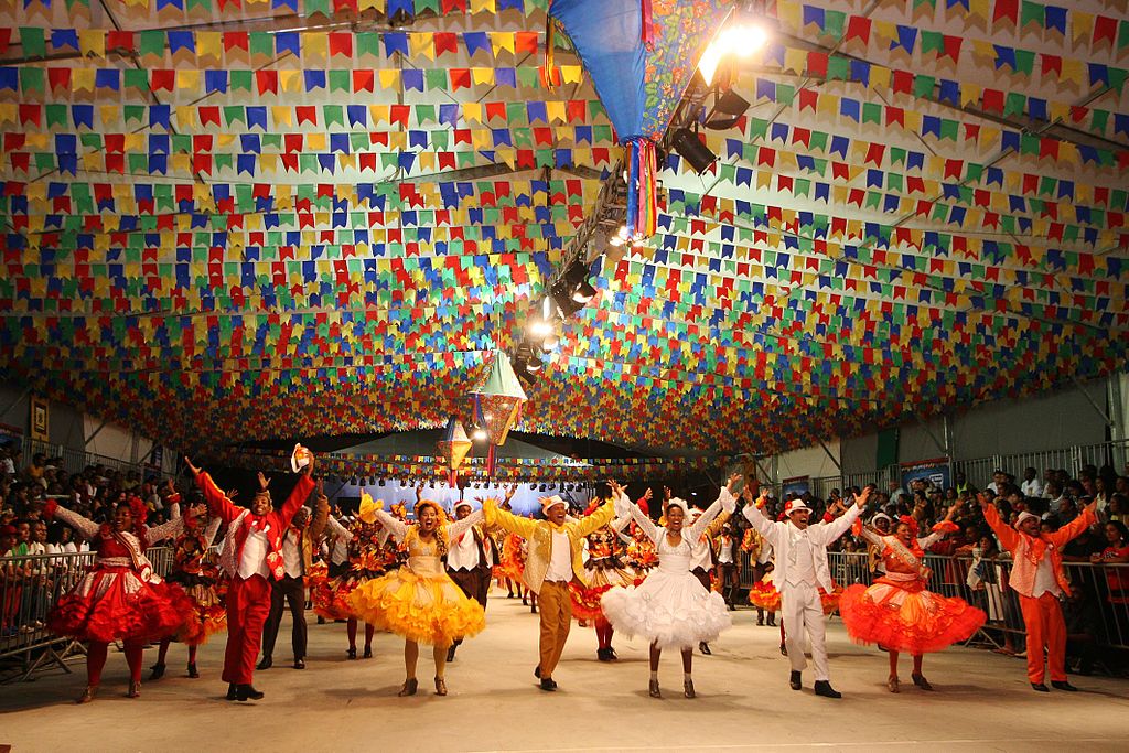 Grupo grande dançando quadrilha em galpão enfeitado para festa junina.
