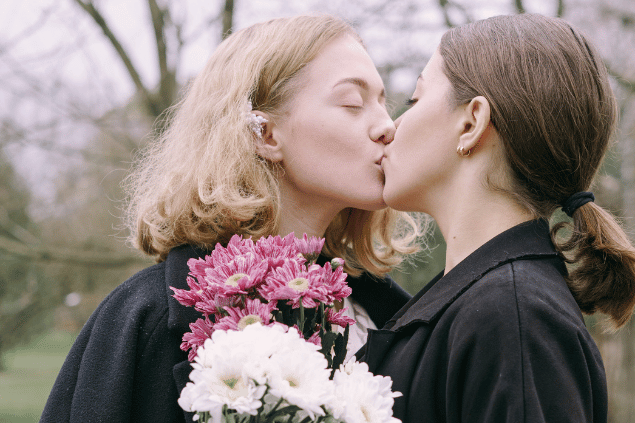 Duas mulheres jovens se beijando enquanto seguram buquês de flores.