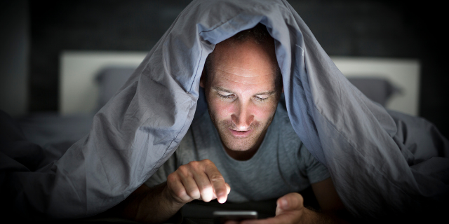 Homem coberto por edredom interage com um celular.
