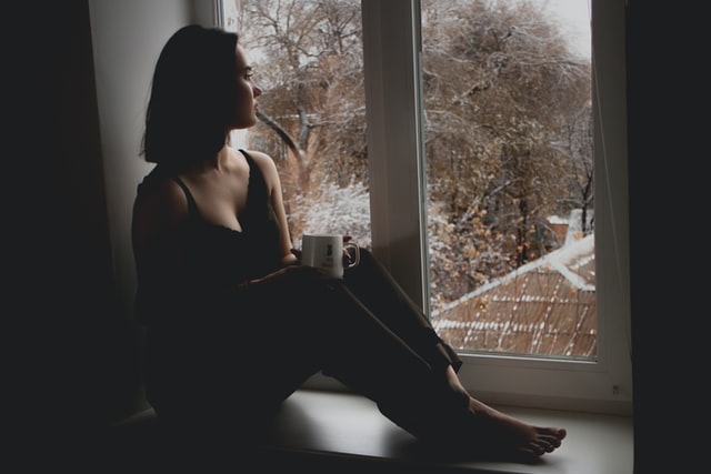 Mulher branca sentada numa janela com caneca na mão.