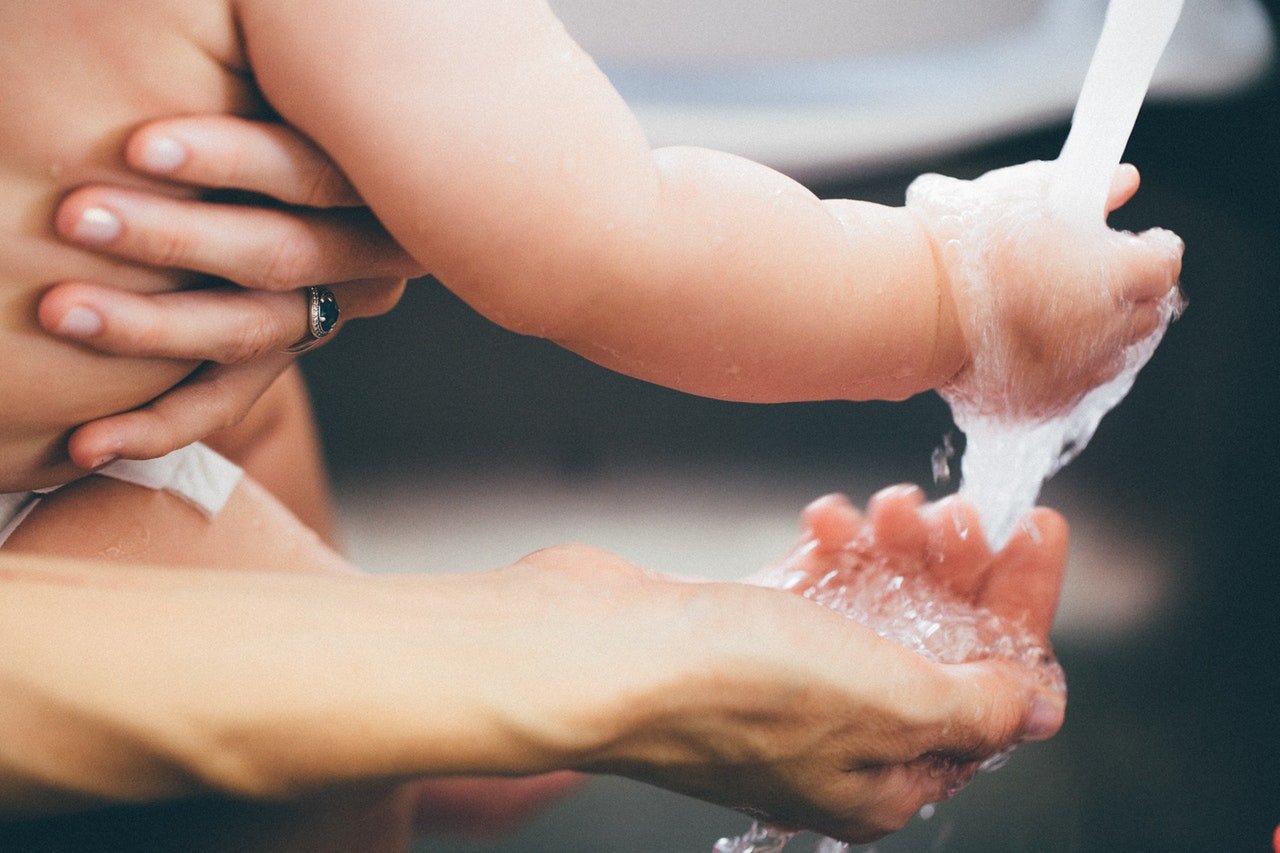 Pessoa segura bebê no colo enquanto os dois colocam as mãos sob uma torneira de água.