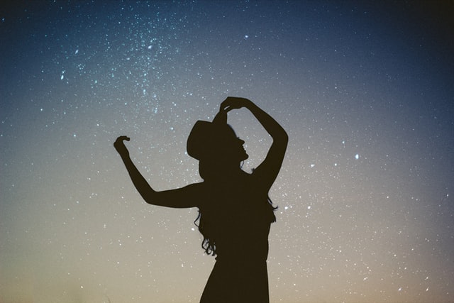 Silhueta de mulher com chapéu e braços levantados com céu estrelado ao fundo.