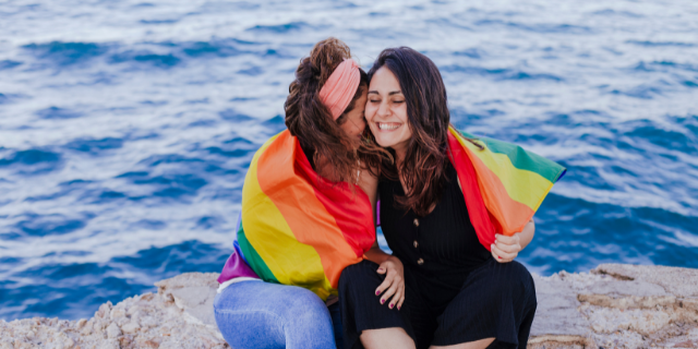 Em praia, casal lésbico abraça-se e sorri. Ambas estão cobertas por uma bandeira representativa da comunidade LGBTQIA+.
