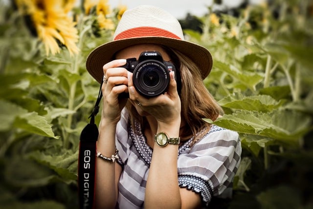 Mulher branca segurando câmera fotográfica num campo de girassóis.