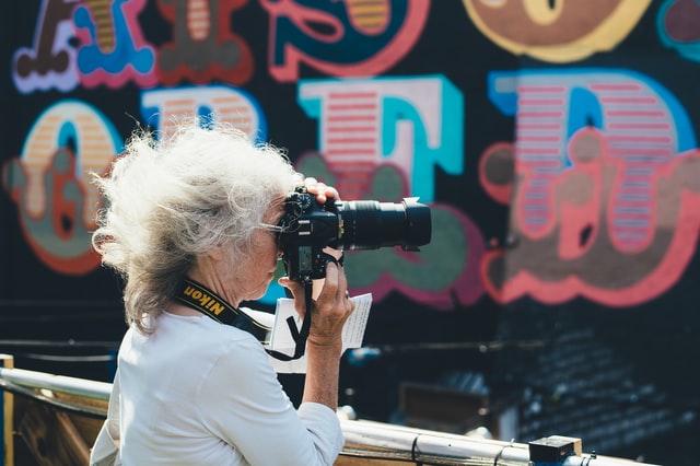 Mulher branca de cabelos brancos segurando máquina fotográfica, com parede colorida ao fundo.
