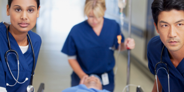 Três profissionais da saúde encaminham um paciente em maca em corredor de hospital.