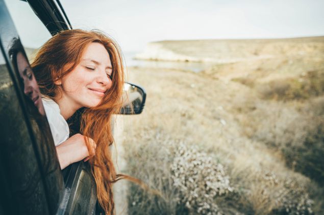 Mariia Korneeva / Shutterstock Mulher sorridente, com os olhos fechados, colocando a cabeça para fora da janela de um carro em movimento.