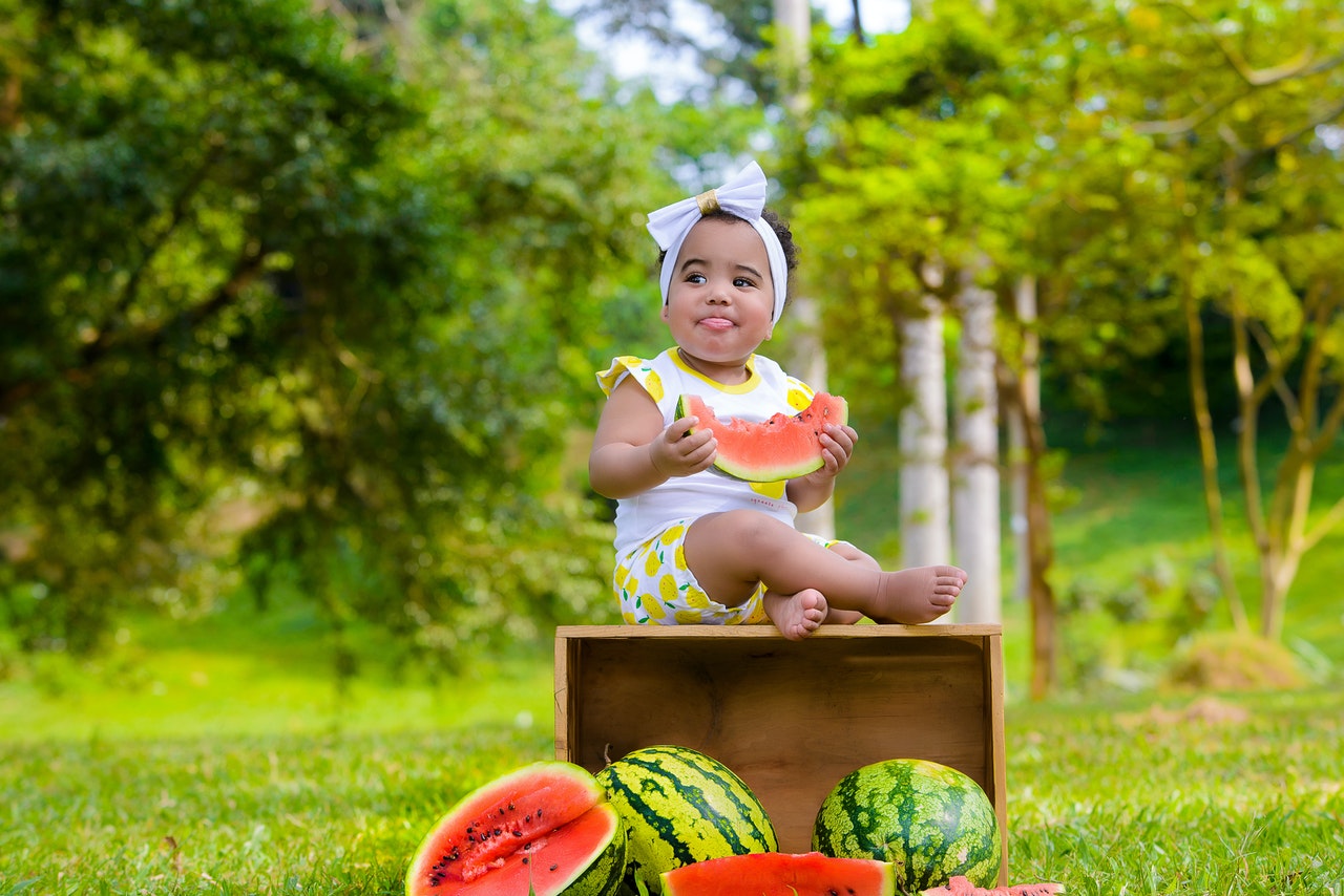 Bebê sentada em um caixote de madeira de lado, que derrubou várias melancias no chão. Ela segura uma fatia de melancia, e lambe os lábios.