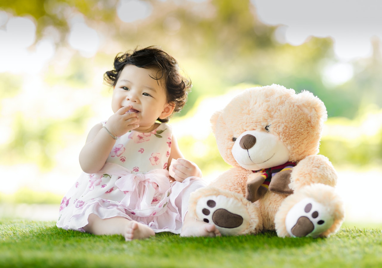 Bebê sentada na grama, com a mão em frente a sua boca, e um ursinho de pelúcia quase do seu tamanho ao seu lado.