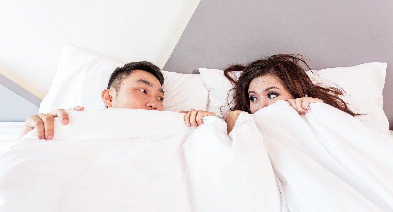 Homem e mulher deitados em uma cama, com o edredom cobrindo metade do seu rosto.