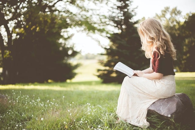 Mulher branca e loira sentada num campo enquanto lê um livro.