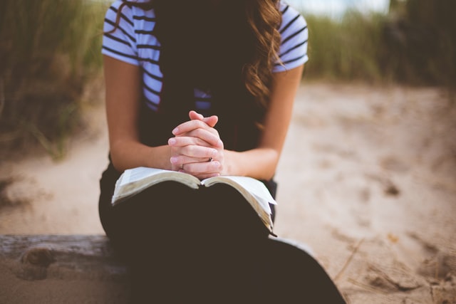 Mulher sentada num banco de madeira com bíblia no colo e mãos entrelaçadas.