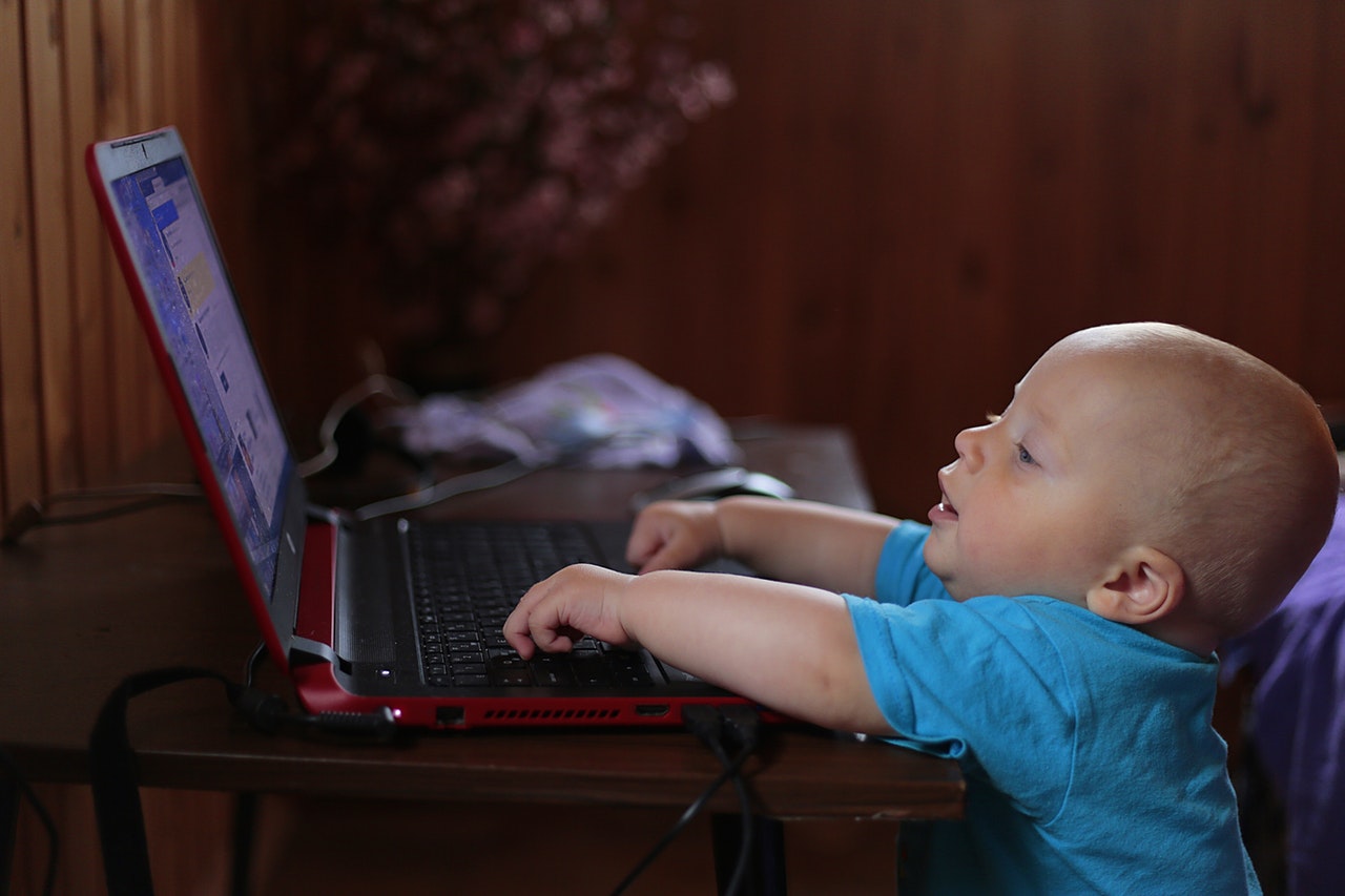 Bebê se esticando para alcançar um notebook em cima de uma mesa, conseguindo colocar as mãos no teclado.
