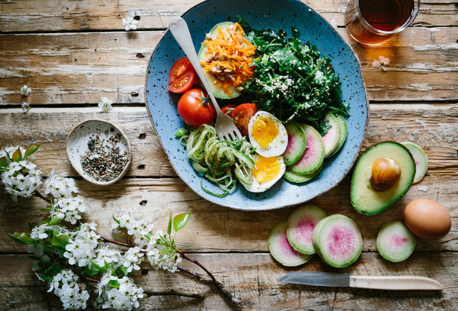 Mesa com prato com diversos alimentos e ao lado um ramo de flor e um abacate cortado ao meio