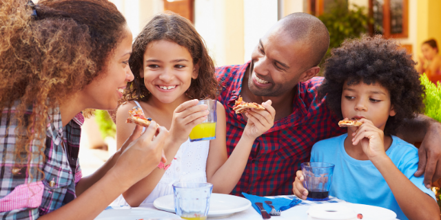 Uma mulher e um homem adultos sentados à mesa junto de duas crianças, um menino e uma menina. Eles comem pizza e sorriem enquanto conversam.