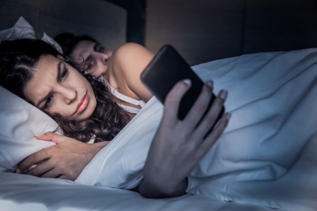 Imagem de um casal na cama, a mulher está olhando no celular no meio da noite de costas para o marido que está tentando ver o que está aparecendo no celular com semblante de desconfiança