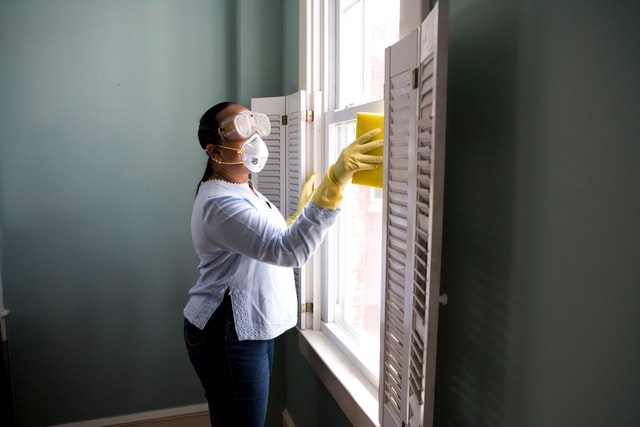 Mulher limpando janela com óculos, máscara e luvas de proteção