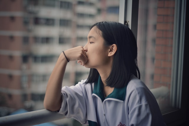 Mulher asiática sentada numa janela com expressão pensativa.