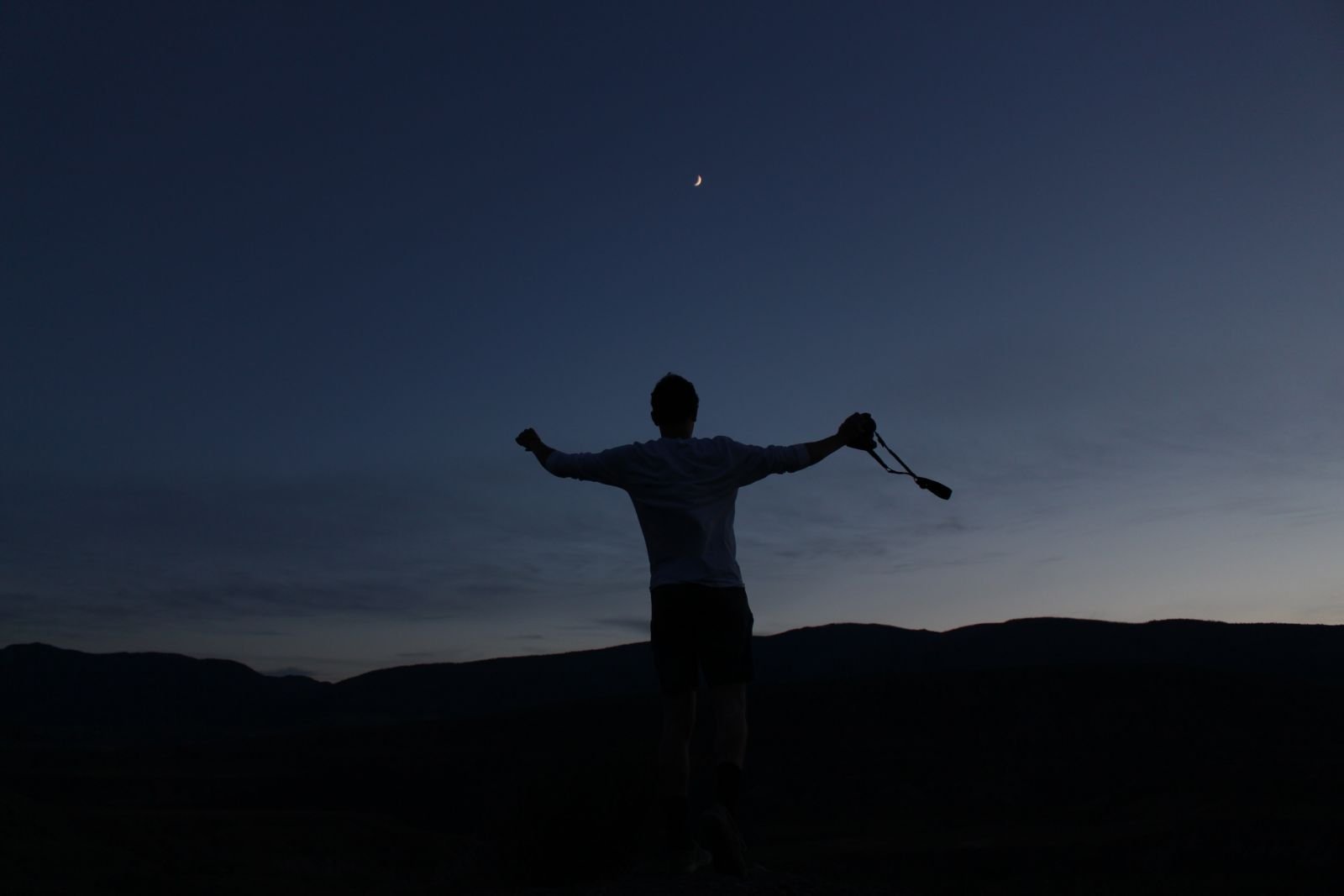 Silhueta de homem com braços abertos segurando uma câmera fotográfica e olhando para a lua no céu
