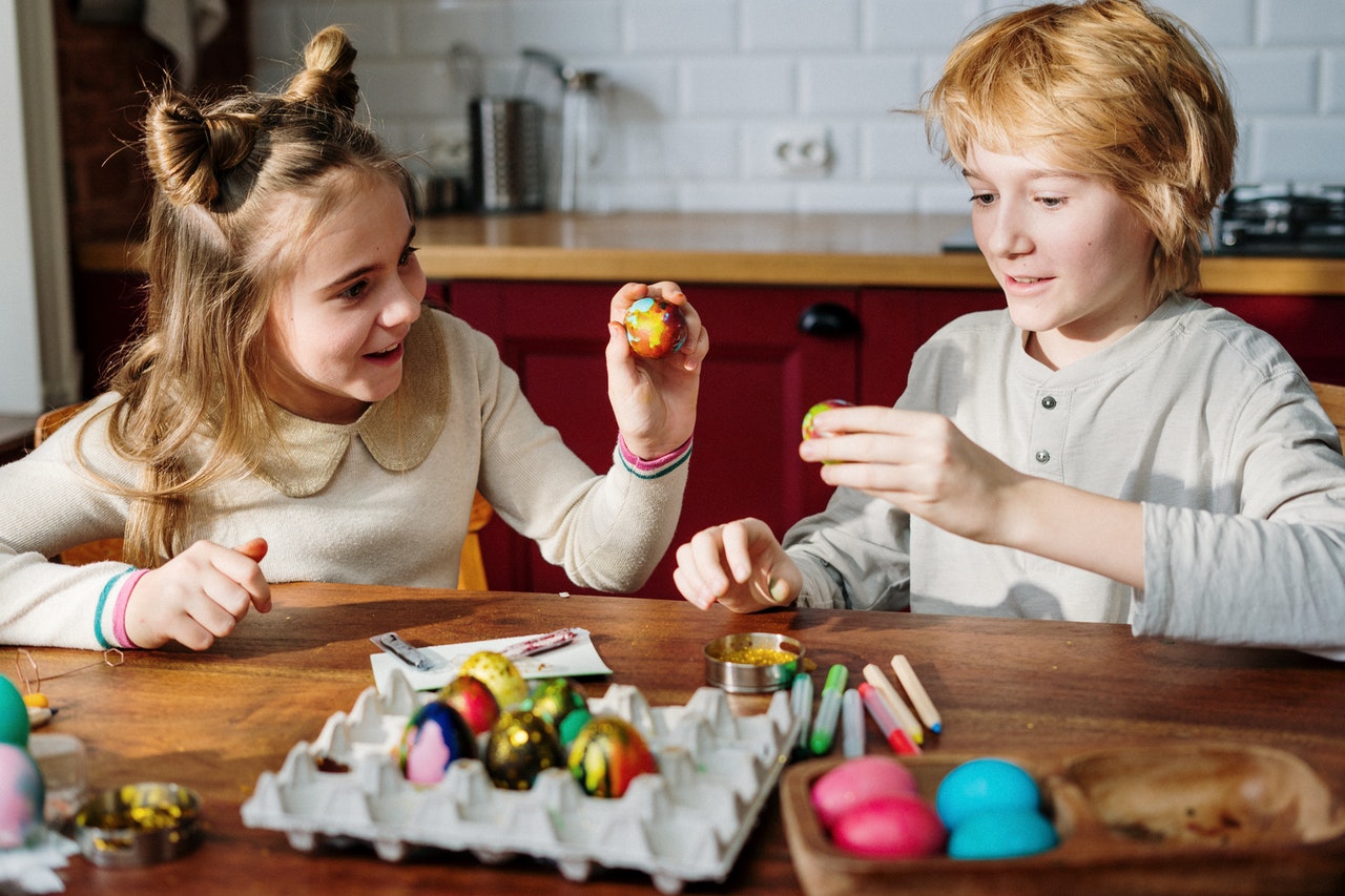 Menino e menina pintando ovos com tintas coloridas. Ambos são loiros e estão sentados lado a lado.