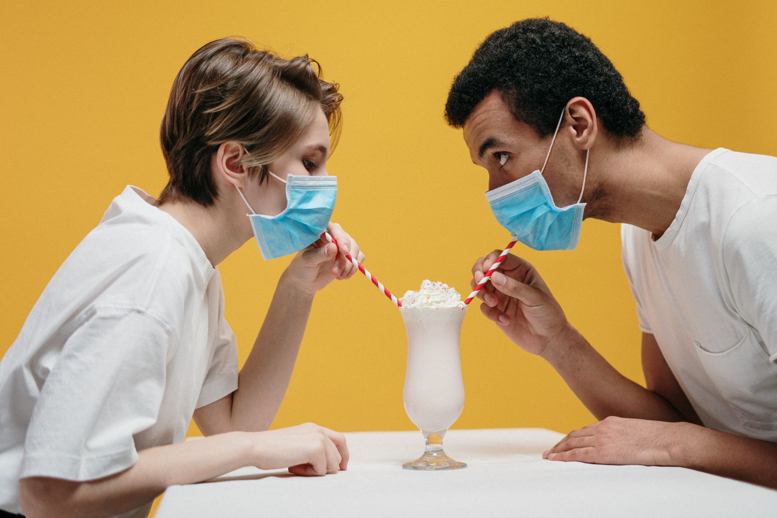 Homem e mulher tomando um milkshake com canudos enquanto usam máscaras de proteção.