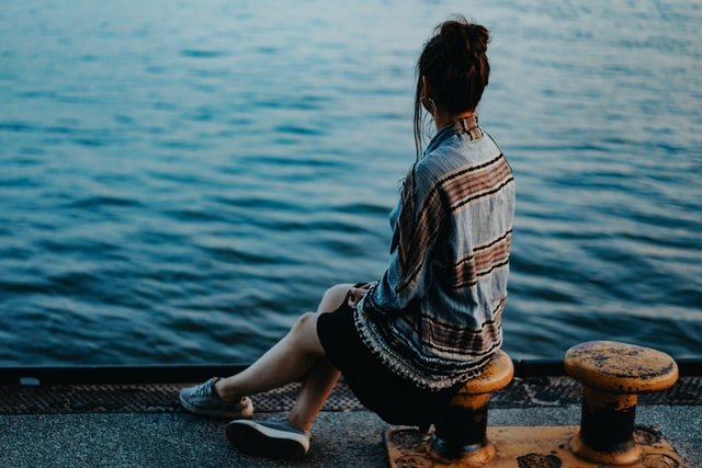 Mulher sentada de costas na beira do mar.