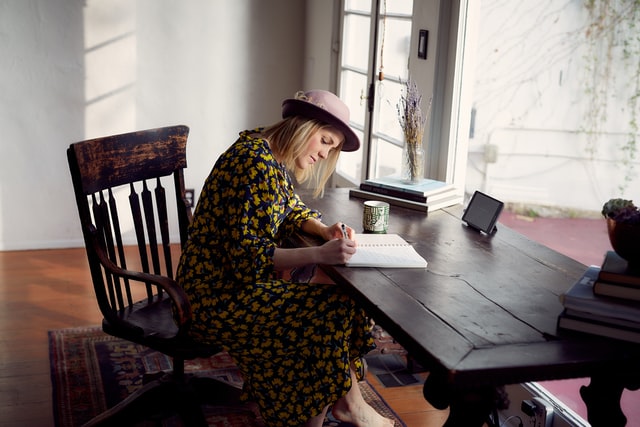 Mulher branca sentada escrevendo em caderno.
