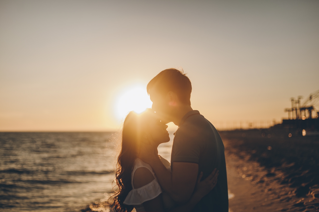 Homem beijando testa da mulher ao lado do mar e do pôr do sol