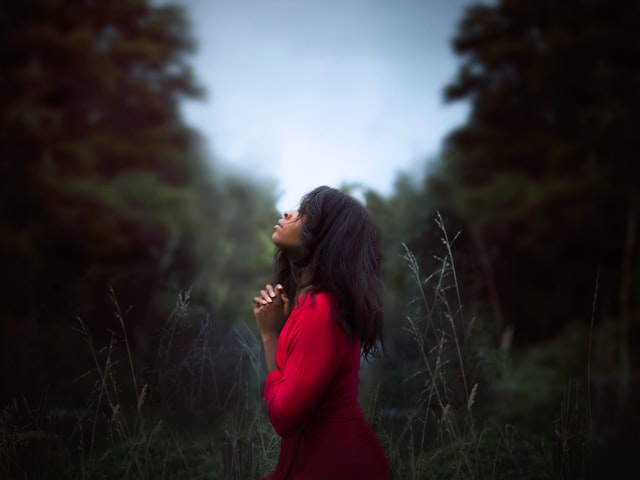 Mulher em oração em pé em campo com árvores ao fundo e céu 