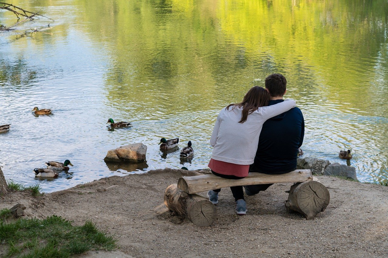 Homem e mulher sentados em banco de madeira, de frente para um lago, observando patos.