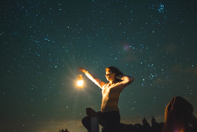 Mulher branca segurando lanterna durante a noite, com céu estrelado atrás.